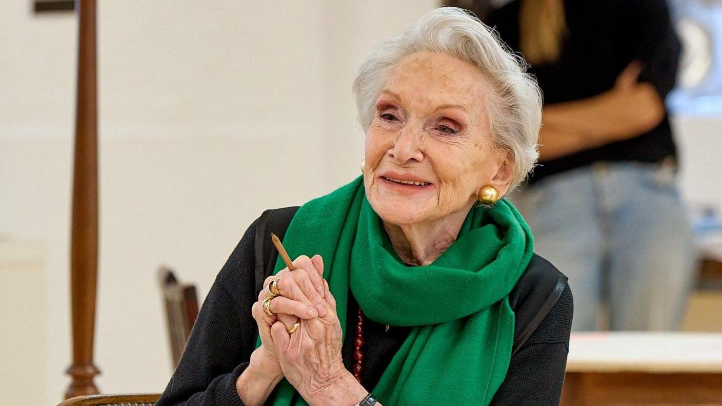 Actress Siân Phillips smiles during rehearsals for Summer 1954, which is being performed at the Bath Theatre Royal. She is wearing a dark top and a long green scarf, and has her hands clasped together