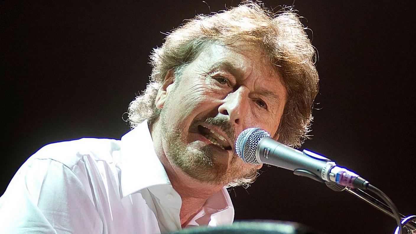 Rick Davies in a white shirt, singing into a microphone on stage at the O2 Arena, London, on October 6, 2010