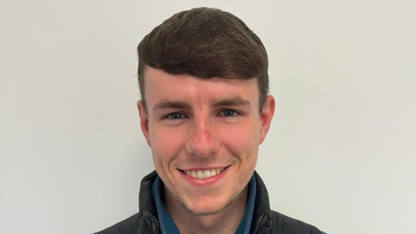 White wall background, shoulders up, Jack is smiling looking directly at the camera. He is wearing a blue fleece under a black waterproof looking coat. He has short brown hair. 