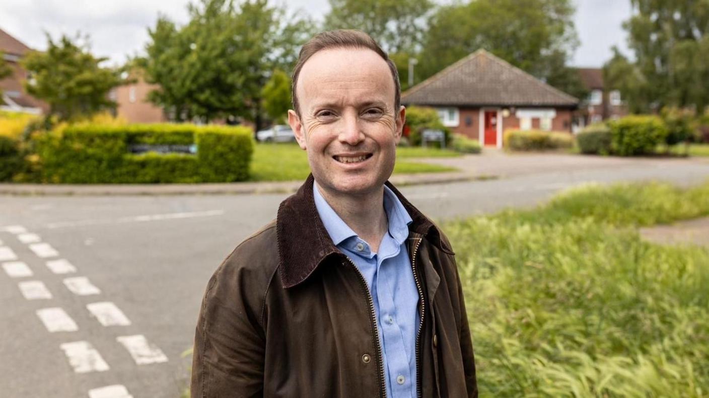 Richard Rout is standing on the corner of a residential street and smiling. He has a brown suede jacket and a light blue button up shirt on.