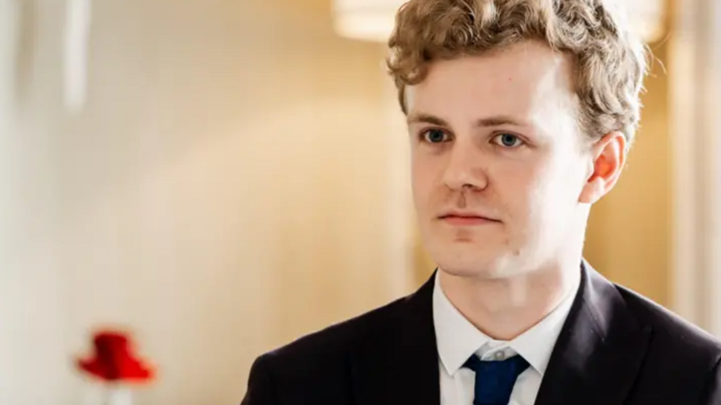 A young man wearing a black suit with a white shirt and a navy tie.