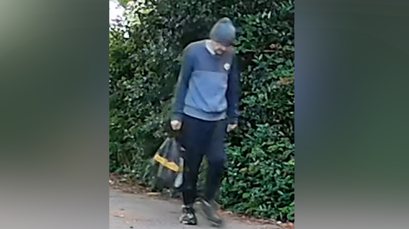 A CCTV image of a man walking on a footpath next to a hedge. He is wearing a beanie hat, a blue jumper and black jogging bottoms and is carrying a bag in one hand. He is looking down at his feet as he walks.