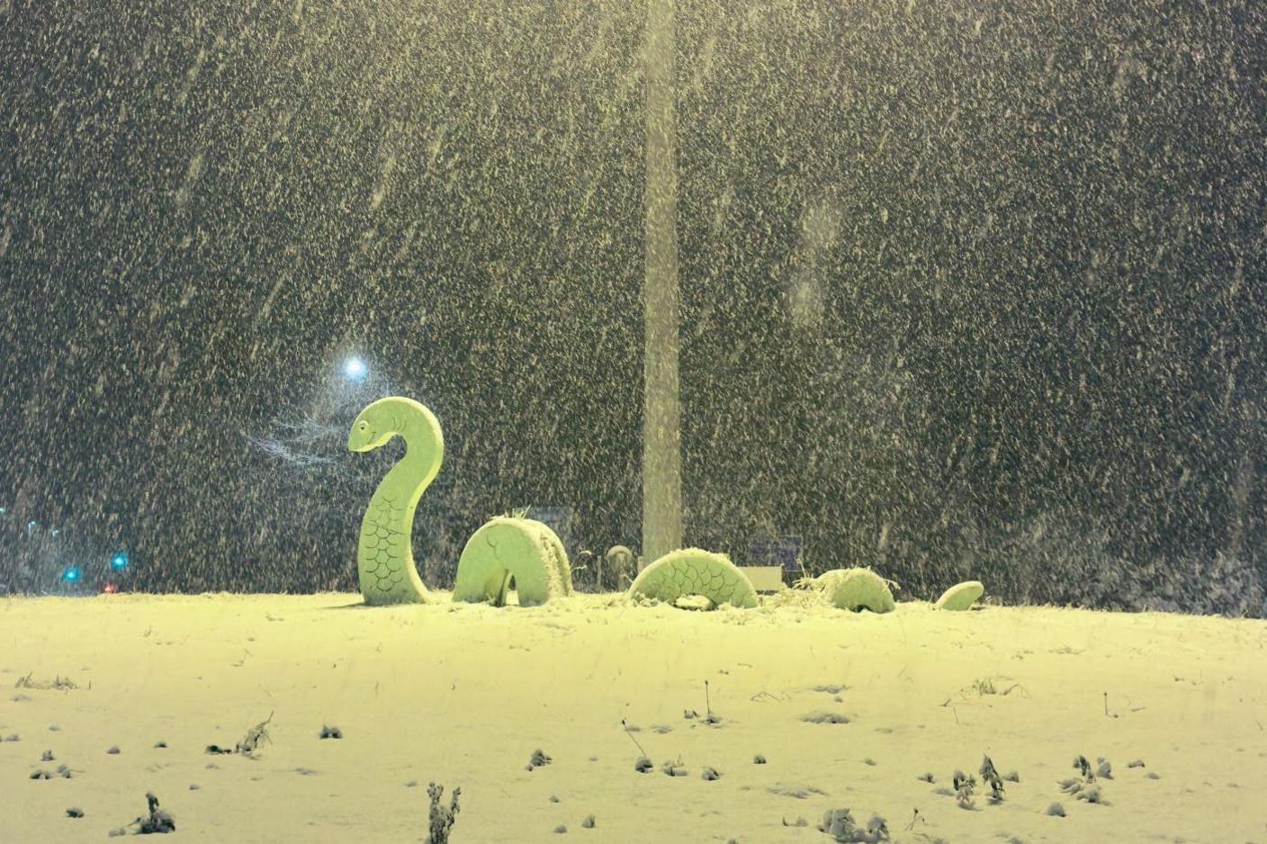 Snow is falling heavily and an artwork of the Loch Ness Monster is illuminated by a streetlight on the roundabout. It is early morning and still dark.