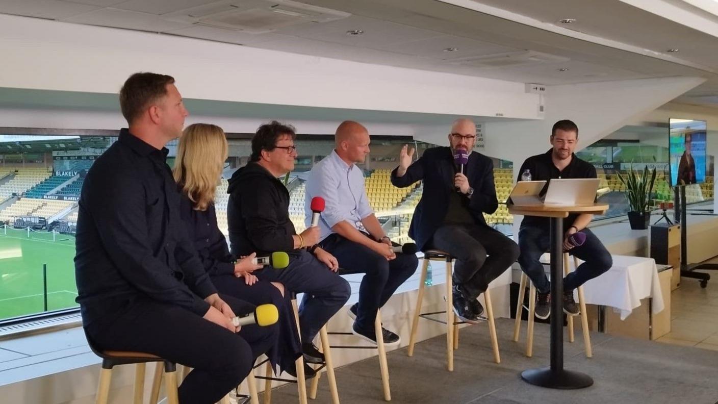Six people – five men and a woman – sitting on tall stools and holding microphones. Behind them, on the other side of a window, is a football pitch.