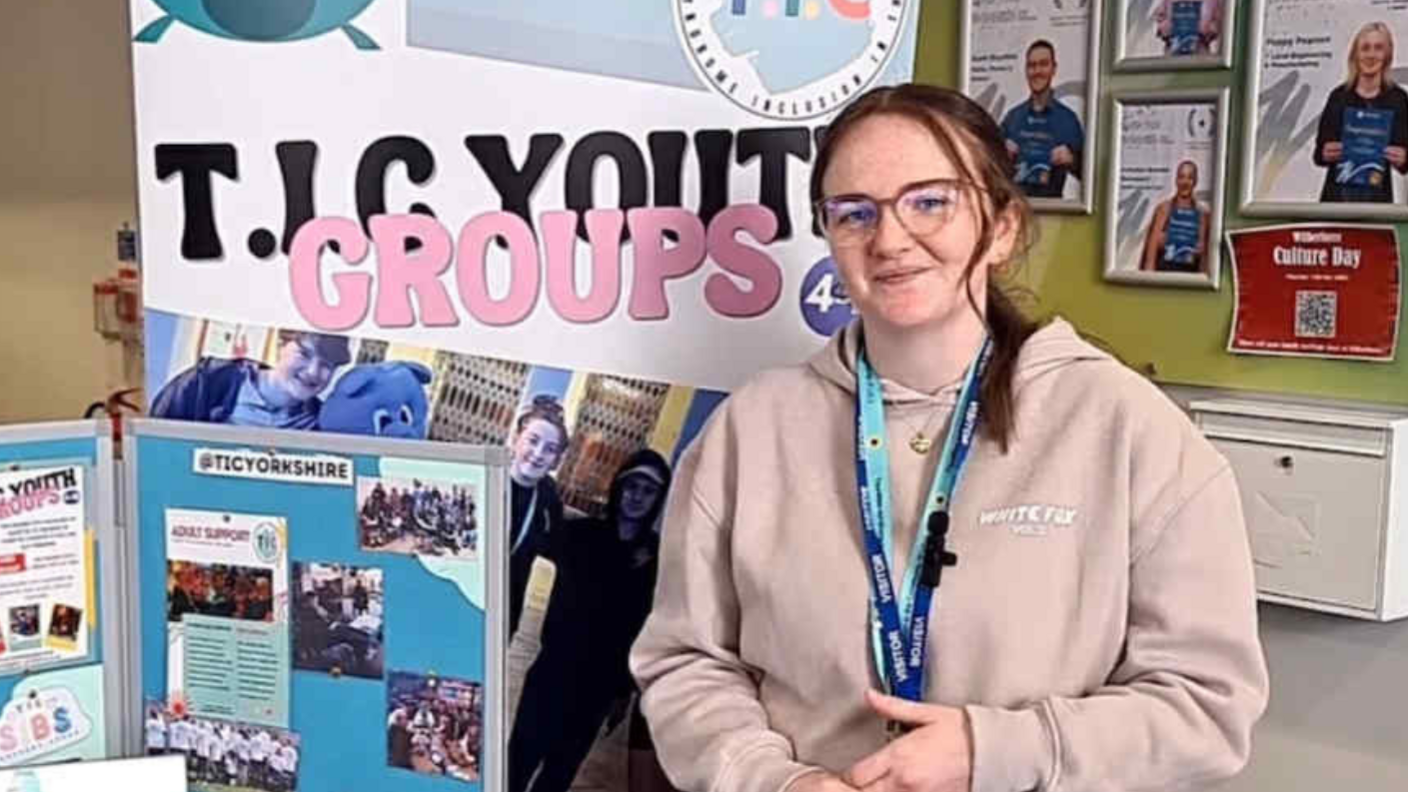 Yasmin Bartle. She has long brown hair and glasses. She is smiling. She has two blue lanyards and a light hoody top. There is a display behind her and next to her with information about Tourette syndrome.