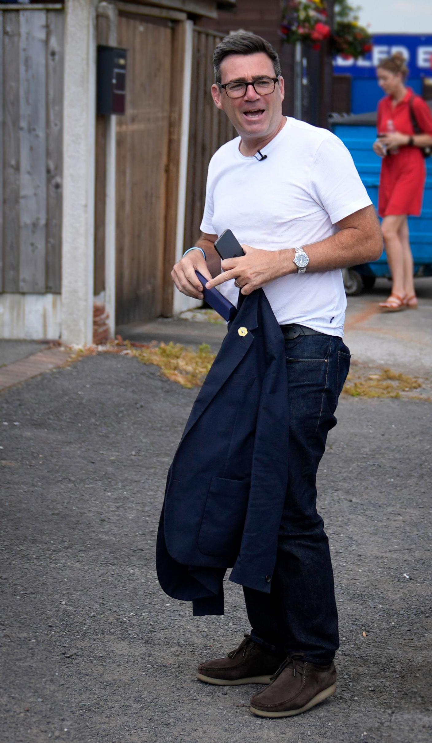 Andy Burnham is walking out of a car wearing a white T-shirt, navy jeans and holding a dark jacket with a yellow badge on the lapel.