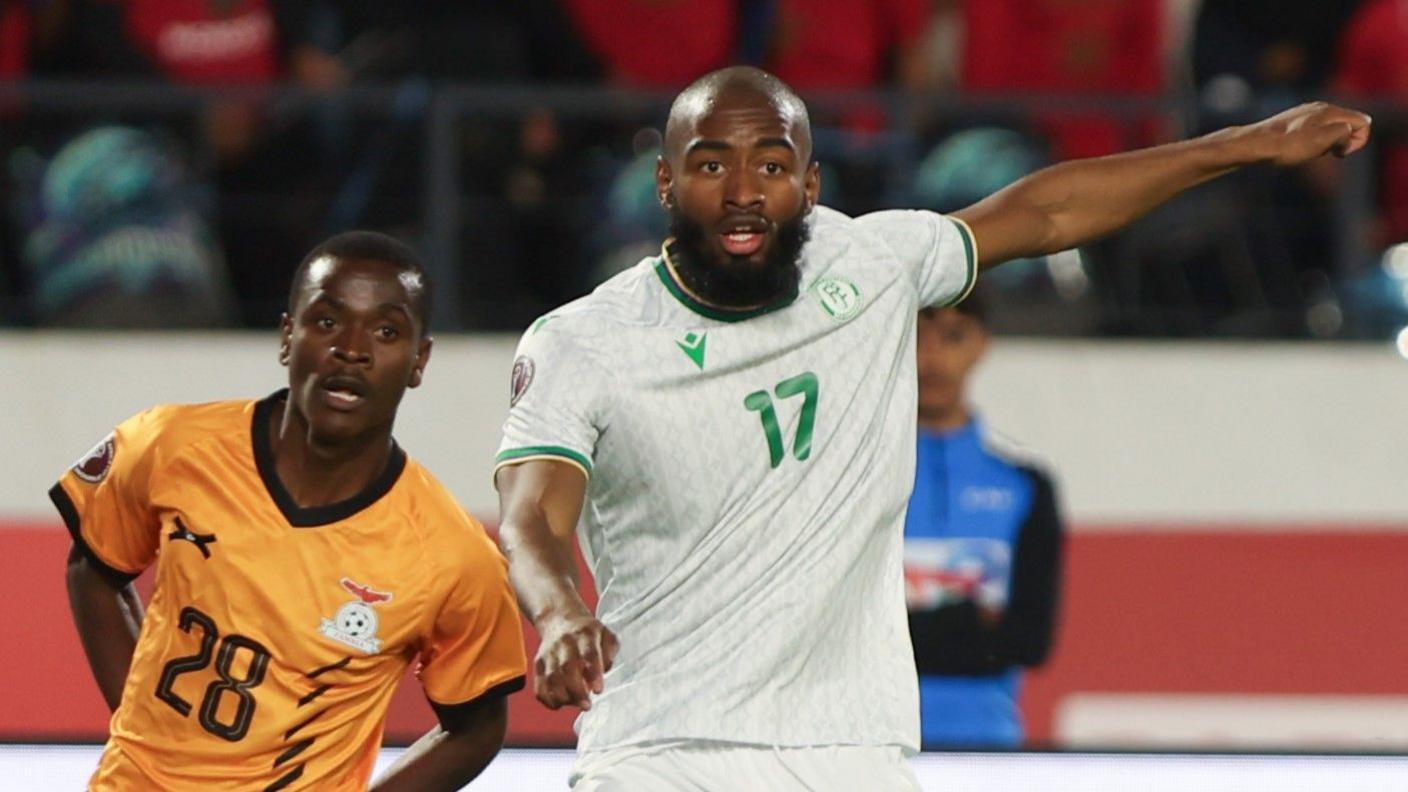 Comoros striker Myziane Maolida (right) and Zambia's David Simukonda (left) seen from waist up during a match at Afcon 2025. Simukonda is wearing a classic gold Zambia home shift and has his hands behind his back, wile Maolida has his left arm extended sideways and his right one slightly raised in front of him, giving the impression of movement. Blurred in the background can be see the high wall of a stand with a ballboy standing in a black and blue top with his arms folded