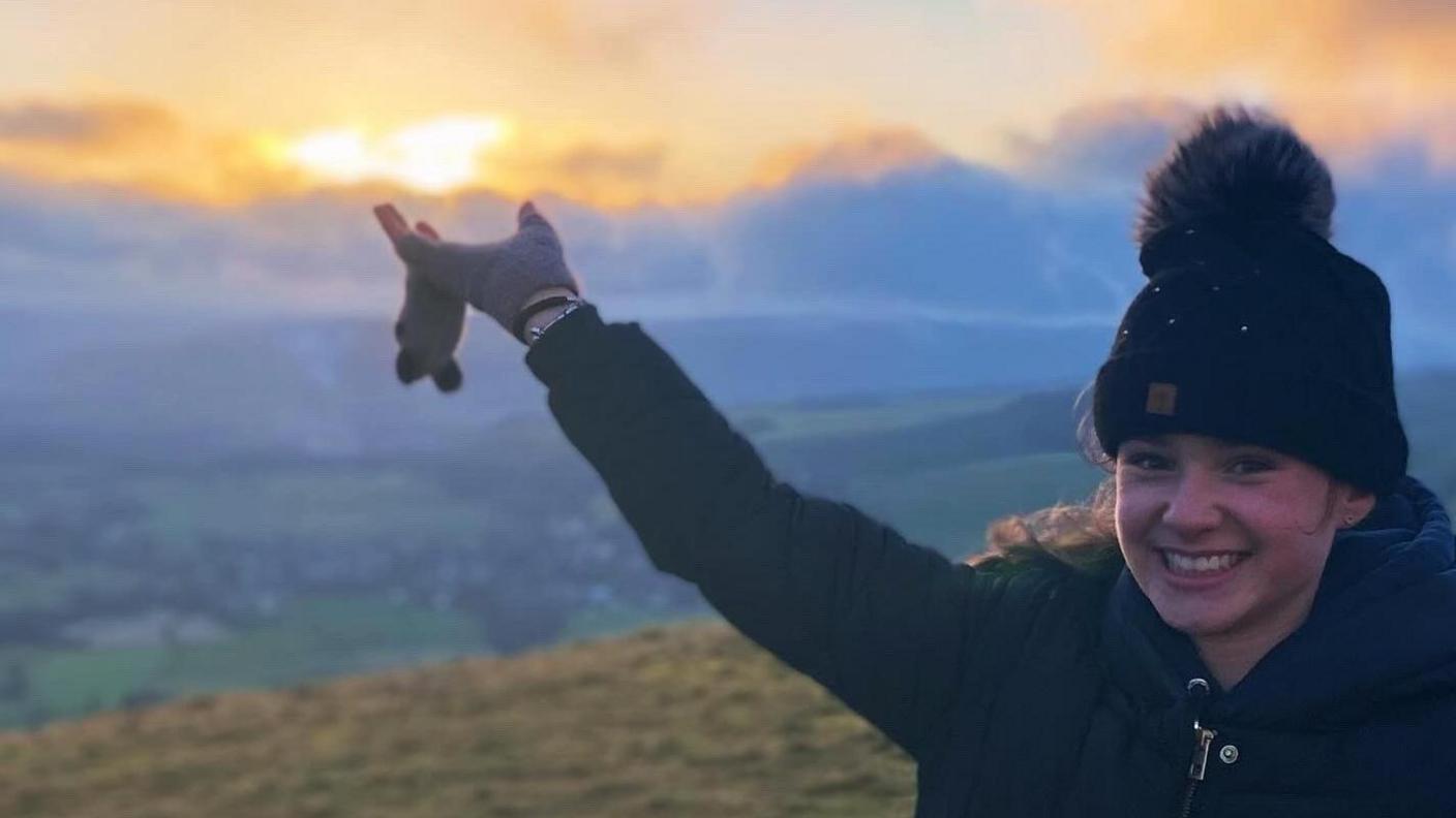 A young girl in a bobble hat smiles as the sun rises behind her