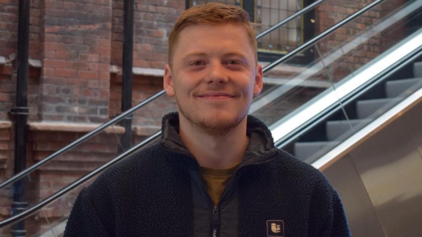 The head and shoulders of a young man, Ben Friel, who is smiling at the camera. He has short red hair and stubble. He's wearing a dark-coloured fleece and green t-shirt underneath. In the background is an escalator and a brick wall. 
