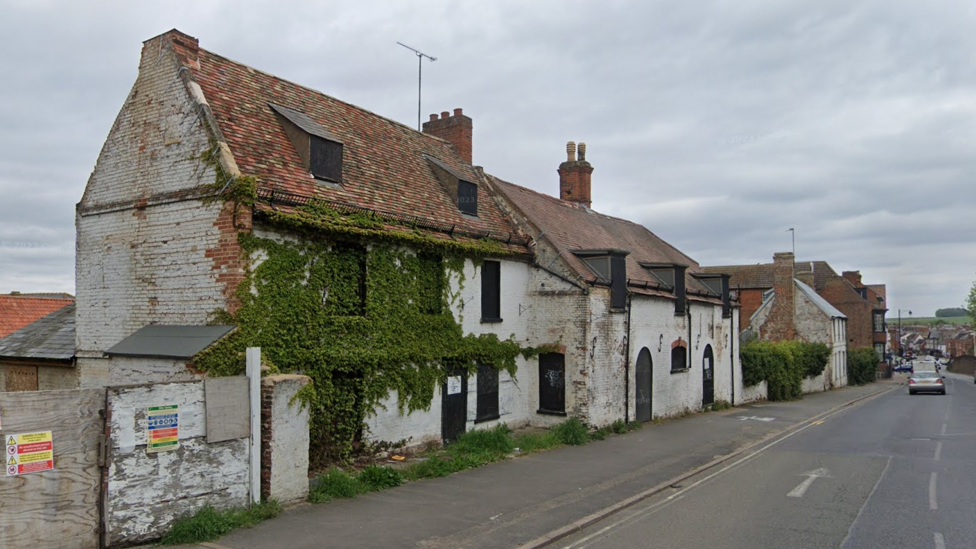 A derelict three-storey building next to a road in Newmarket. It is overgrown with vegetation and weeds and looks in a poor condition.