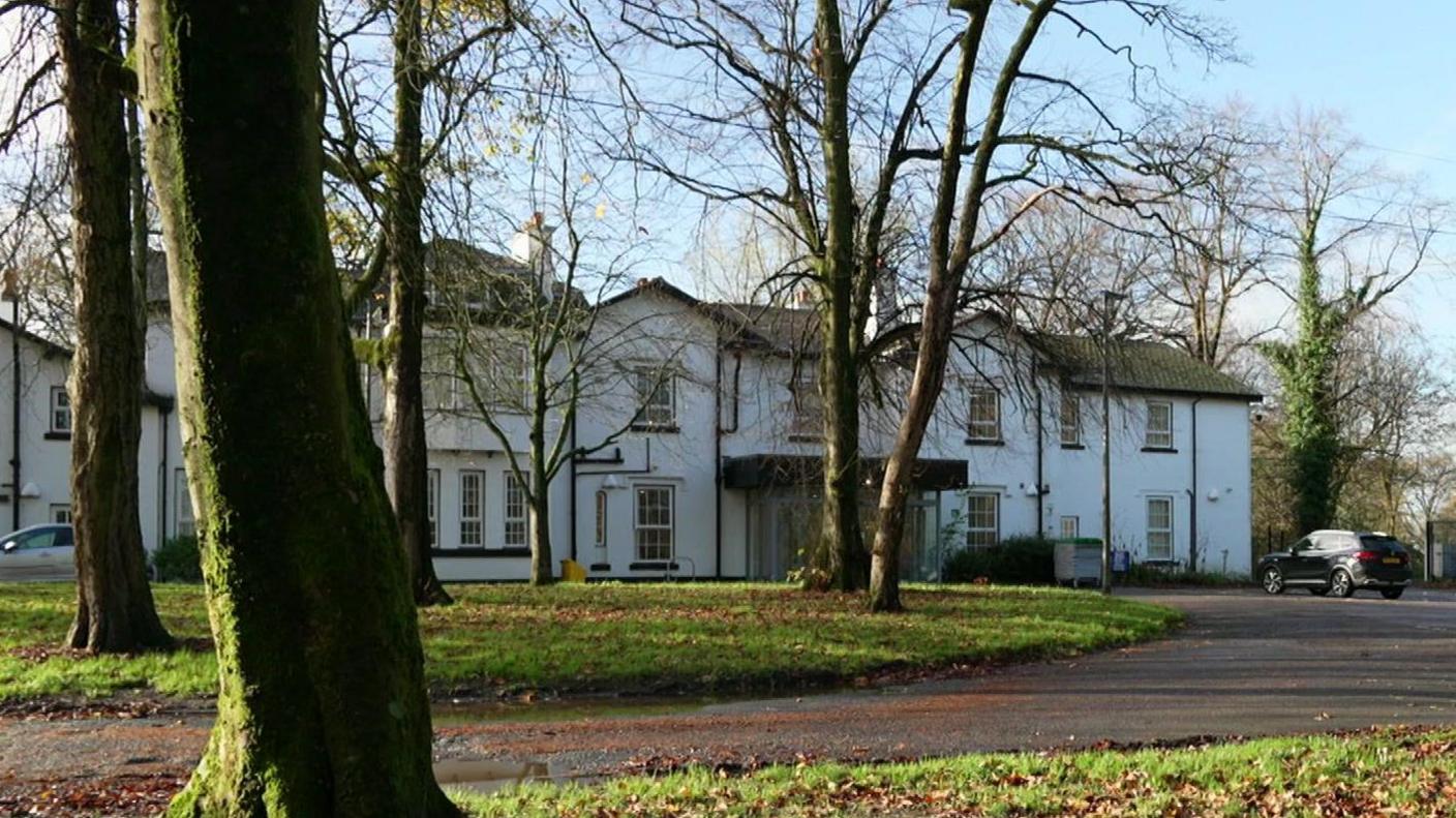 A white building set back behind trees with a car park in front