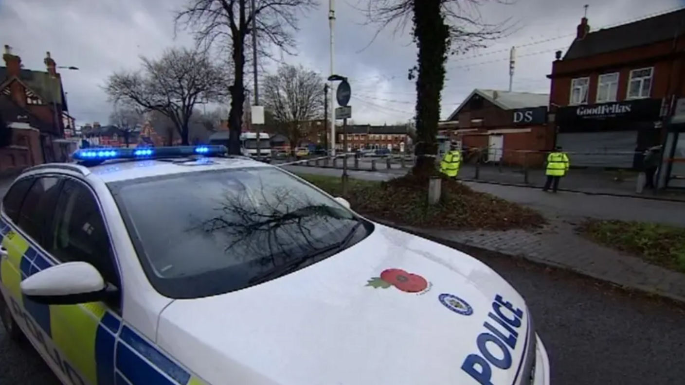 A white police car with a poppy and the word Police on the bonnet, parked in a dark street with houses in the background and two police officers with reflective jackets