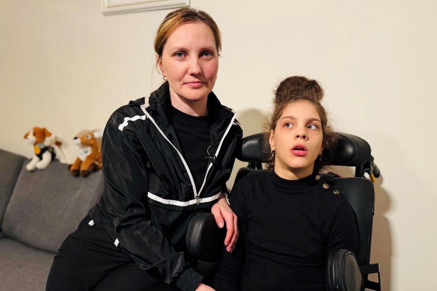 Mother and daughter Olha and Inna Karpets, both dressed in black, the teenage girl is in wheelchair and her mother is smiling at the camera, her hand resting on her daughter. There are soft toys resting on the back of the sofa behind them.