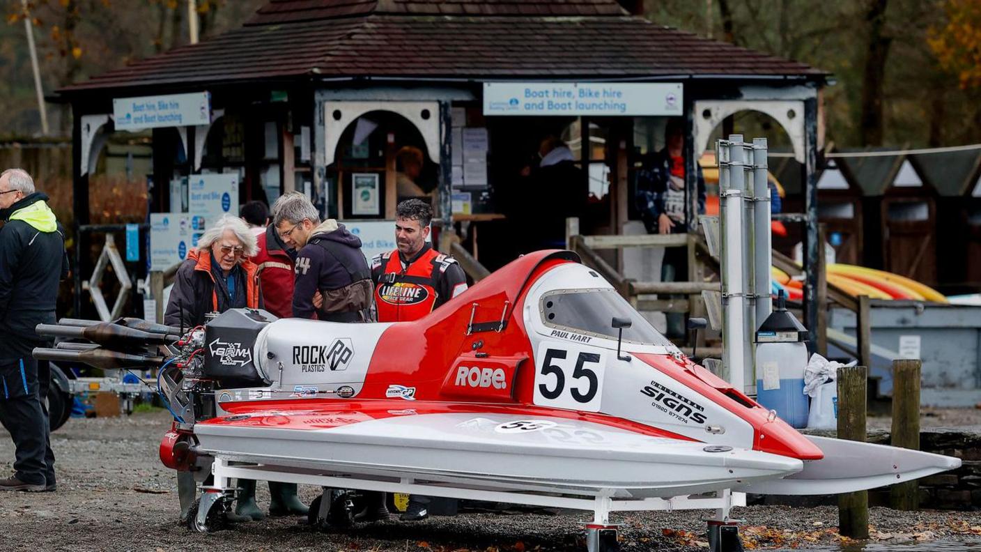 Red and white powerboat with the number 55 written on the side. It is sitting on the edge of the lake. There are four people just behind the boat. Two are looking at the back of the boat which has a curved frame and wings.