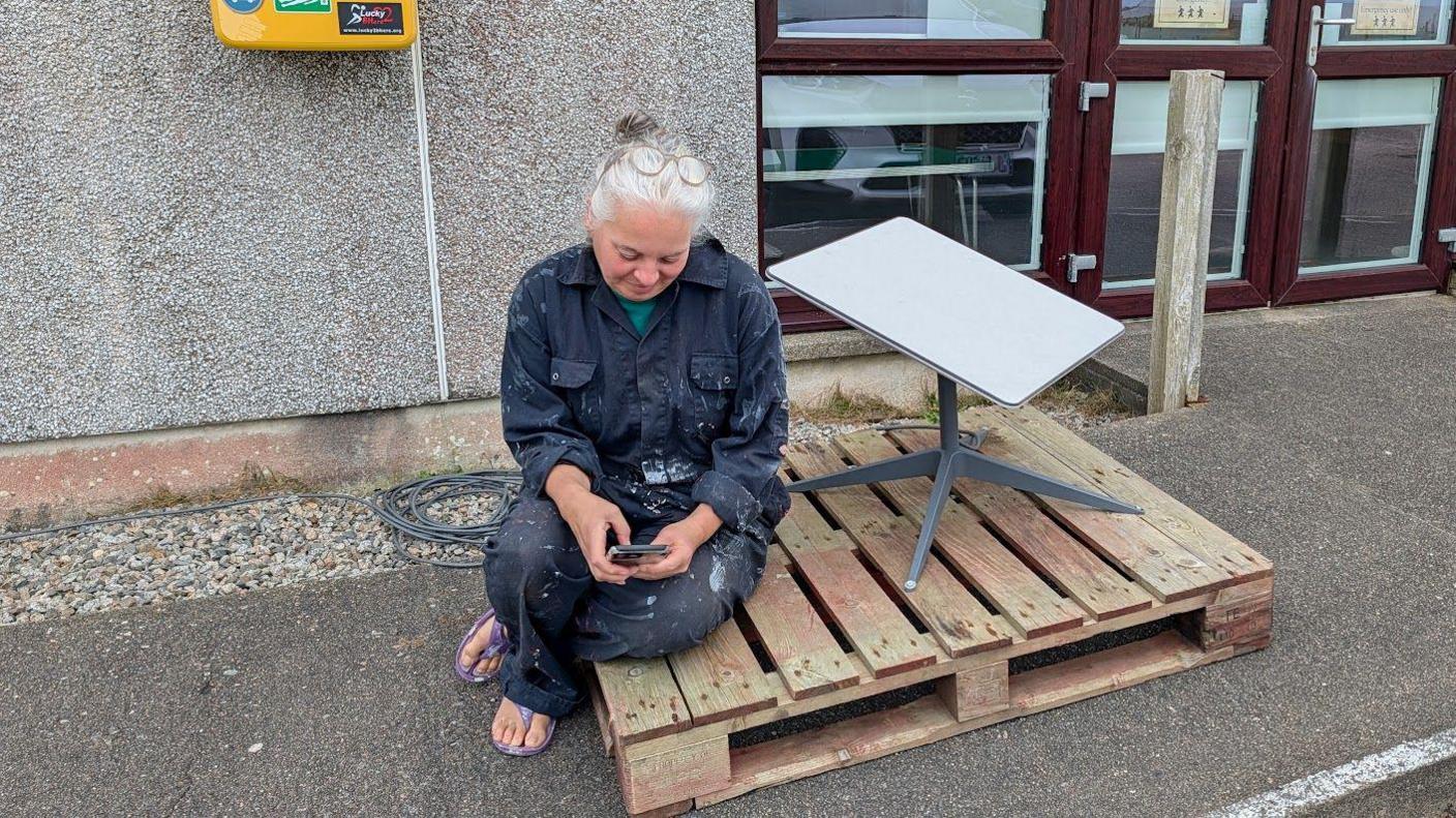 A woman sitting on a pallet in front of a building, while looking at her phone.