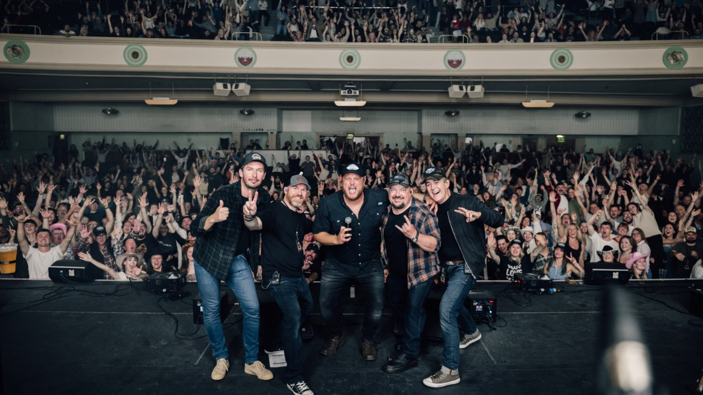 The five band members are standing on a stage with their backs to the audience who are cheering and waving in the background.