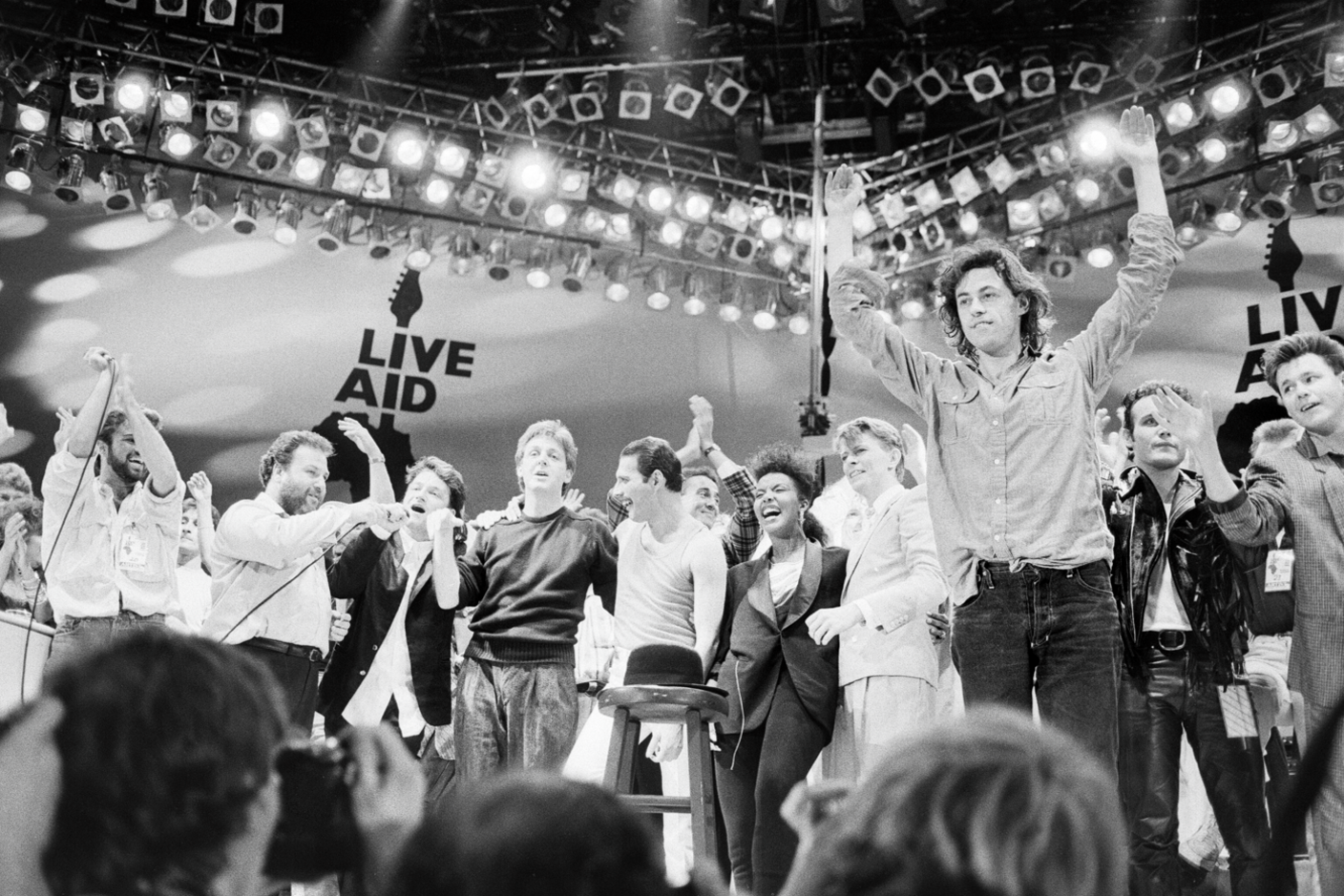 Black and white photo of various stars who performed at Live Aid on the stage including Bono, Paul McCartney, Freddie Mercury, David Bowie and Bob Geldof