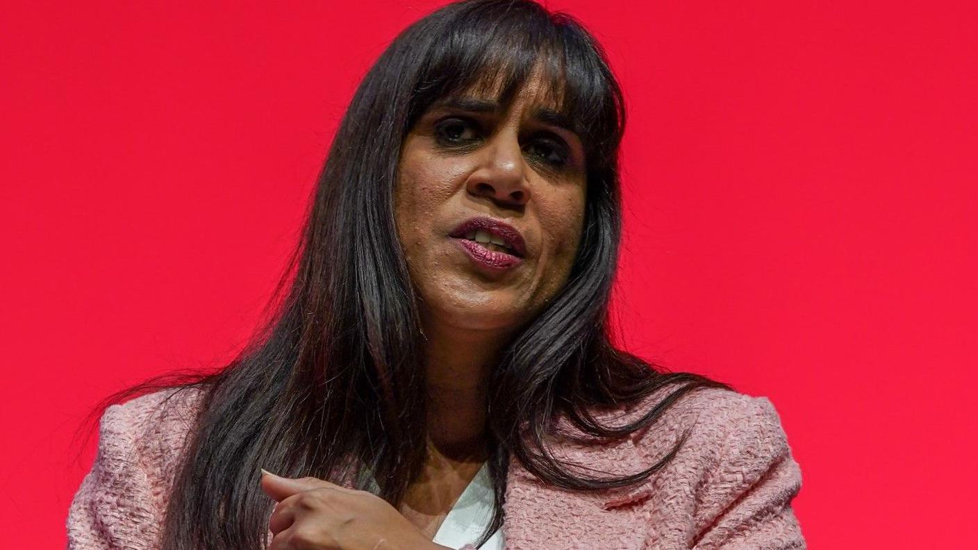 A woman with long dark hair is wearing a pink jacket. She is in front of a red background.