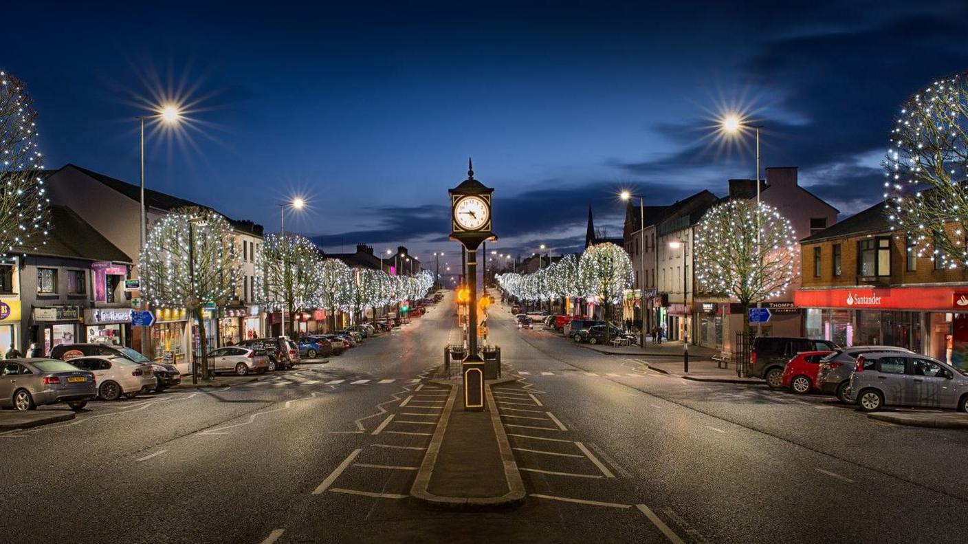 A two way street with trees that are lit up on either side. Shop fronts and cars are along the side. A clock tower is in the centre of the frame, in the middle of the road. 