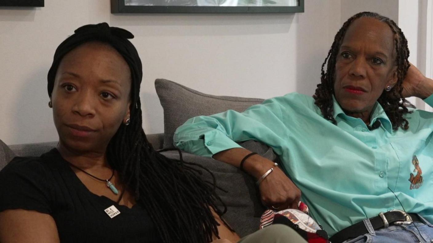 TJ Jacobs, wearing a black t-shirt and a necklace, is sitting on a grey sofa next to Vyleen Maximen, who is wearing a mint green shirt, black belt and blue jeans