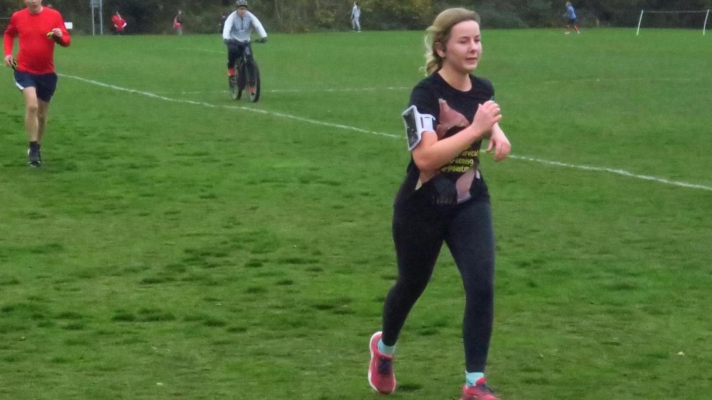 Mrs Lawrence is pictured running during the Kesgrave Parkrun on a football pitch. Other runners can be seen behind her as well as a person riding a bicycle. 