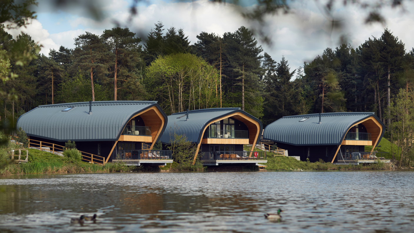 Three wooden waterside lodges with grey roofs are shown with a lake in the foreground and trees in the background