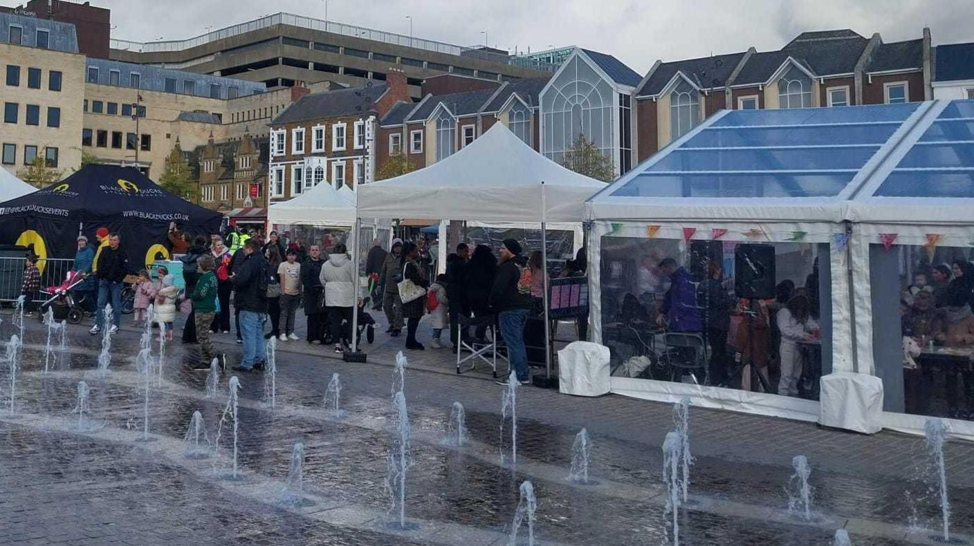 A market square with lots of people waiting in and outside of multiple gazebos. A fountain in the middle of the square is firing water up.