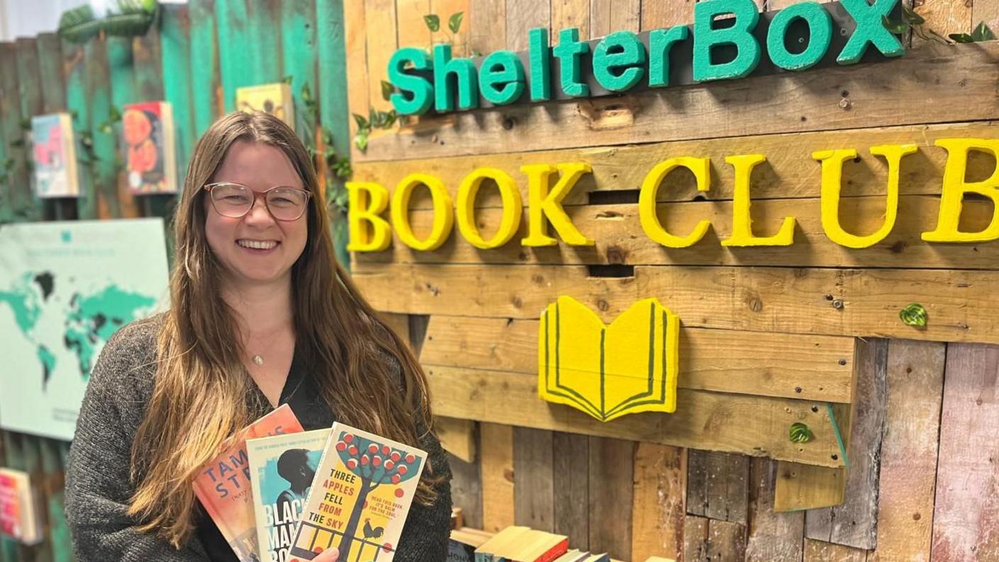 A smiling woman holds some books and stands in front of more books, an the words ShelterBox and Book Club.