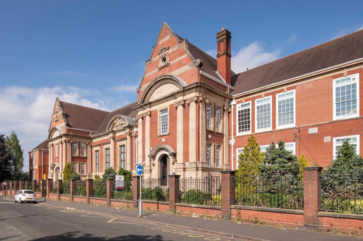 A red brick school with chimney and white windows with railings surrounding it.