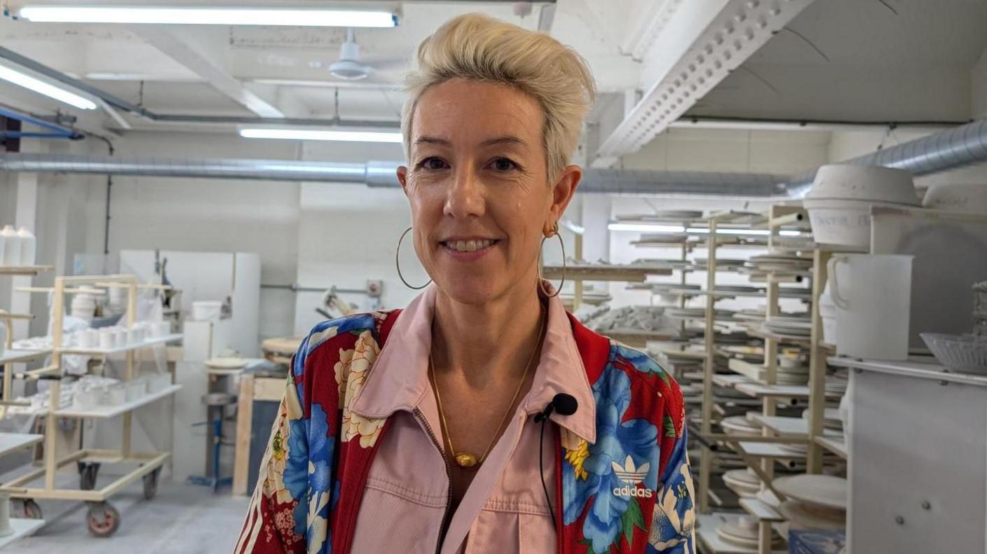 Emily wears a colourful jacket over pink overalls, she is wearing hoop earrings and has bright blonde, short hair with a quiff. She is on the factory floor surrounded by shelves displaying mugs and plates, and looking directly at the camera.