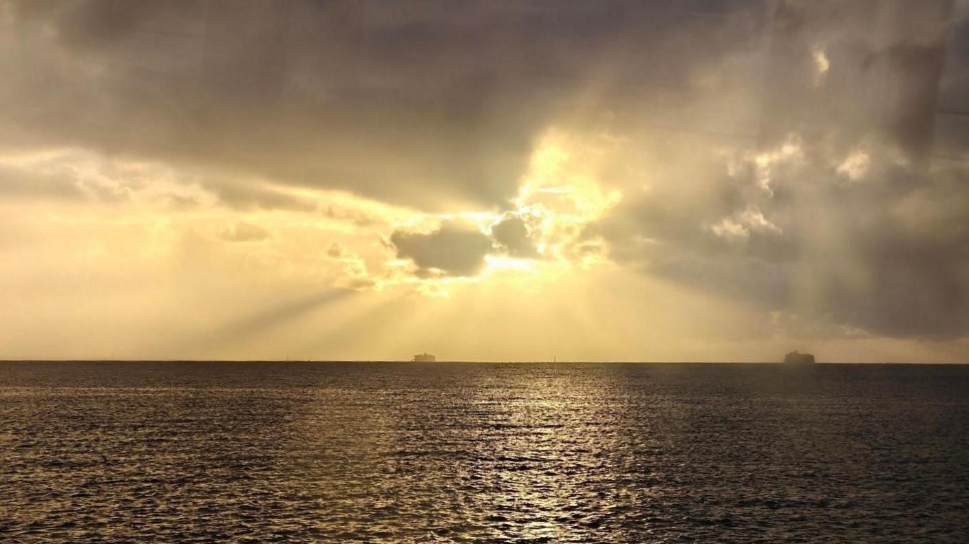A grey sea reflects yellow sunlight across it's choppy surface. The sun is hidden behind an equally grey cloud with beams of golden sunlight emanating across the sky