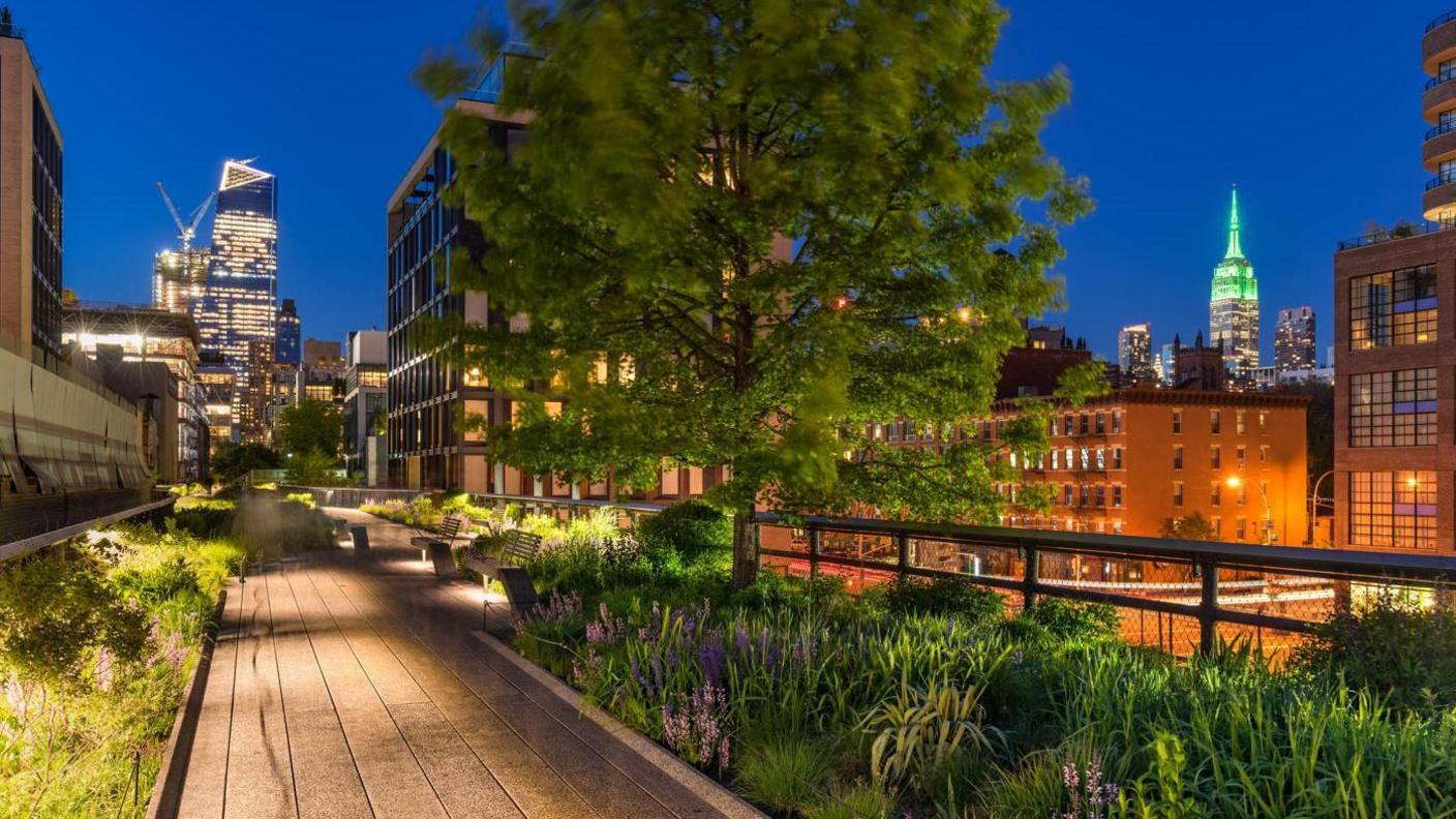 A well-lit walkway is shown at dusk, with trees and buildings to the right and in the background 