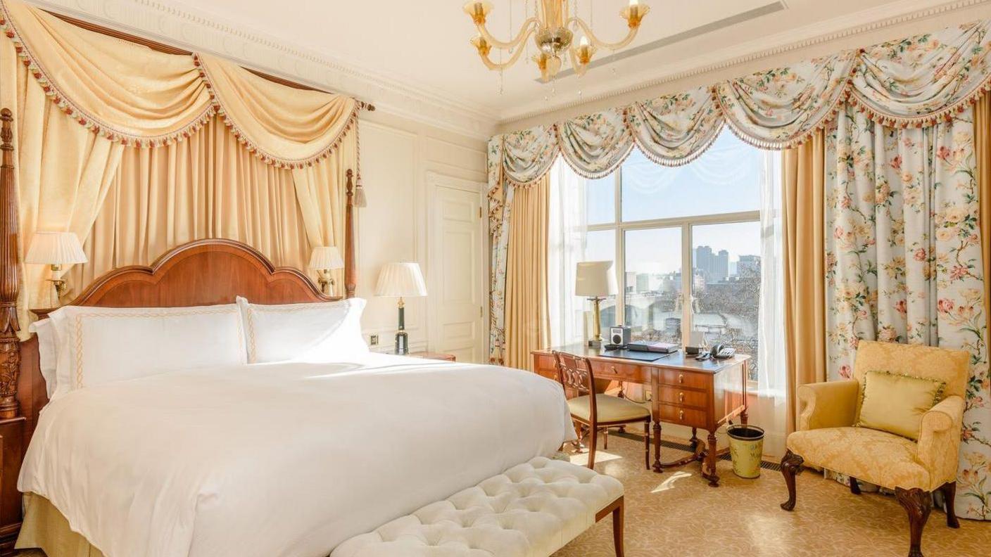 A guest room inside The Savoy Hotel. Floral drapes are across a window. There is a desk and seat facing out of the window. A yellow armchair is in the corner of the room. A large wooden bed has cream drapes behind it. 