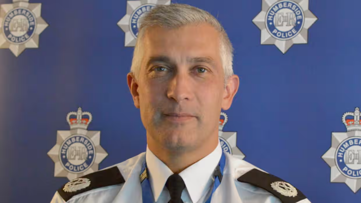 A head and shoulders image of Paul Anderson, the former Chief Constable of Humberside Police. He has grey hair and is wearing uniform consisting of a black tie, white shirt and police lapel badges. He is standing in front of a blue wall featuring multiple Humberside Police logos.