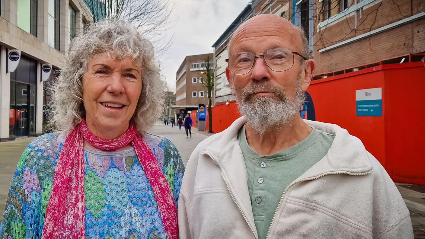 Valerie and Chris standing together on a street in Swansea. She is wearing a brightly coloured crochet top and pink scarf. He is wearing a beige fleece jumper and green button down shirt. 