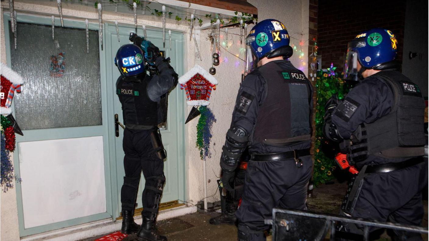 A police officer dressed all in black wearing a blue helmet from a tactical squad cuts open a sky blue door at a home covered in Christmas decorations. Two other officers wait behind him to enter. 