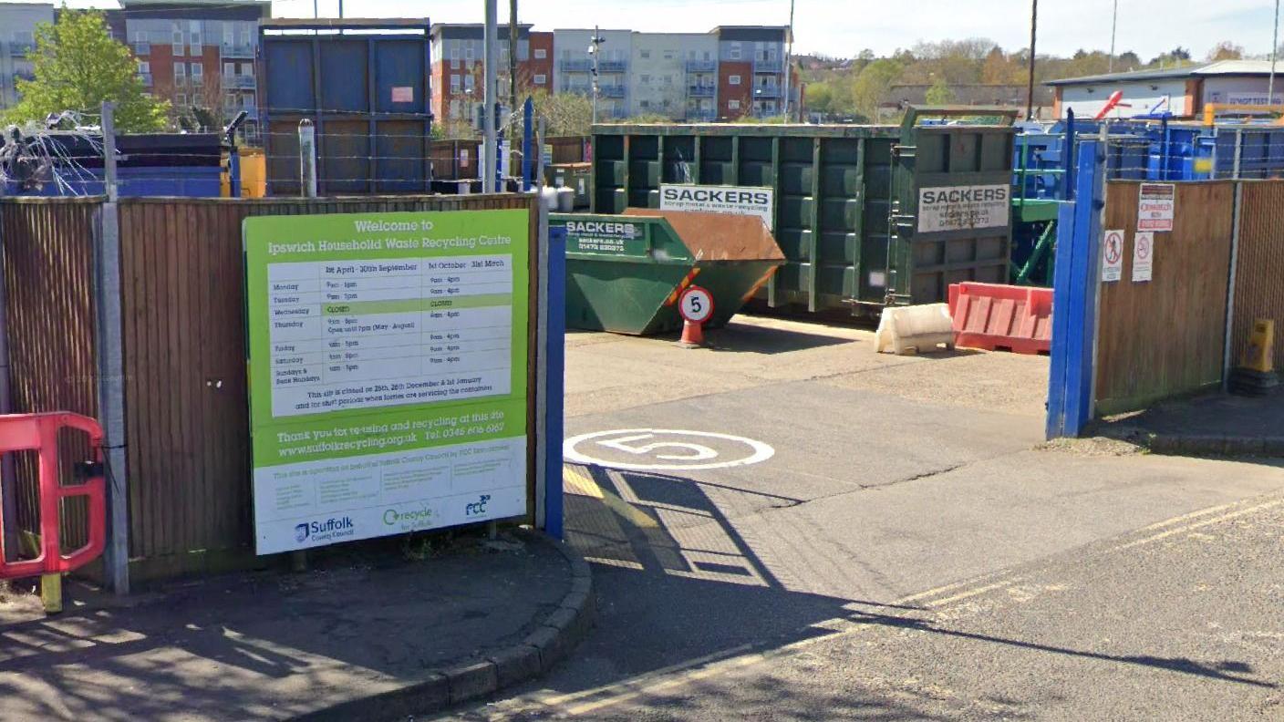 The entrance of a household waste recycling centre with a skip and large mental containers close to the entrance, wooden fences and a tarmac surface 