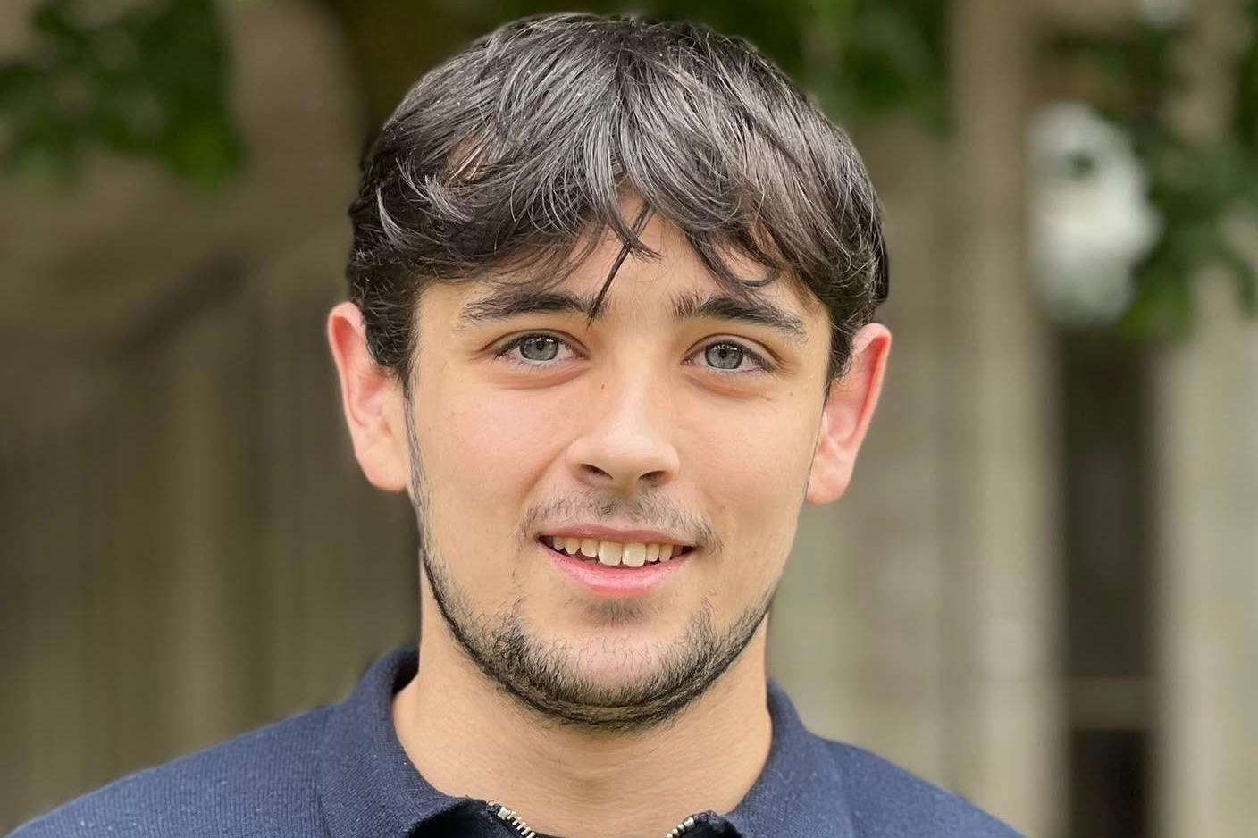 A young man smiles at the camera, he has dark brown hair and facial hair, and blue eyes. He is wearing a navy jumper. 