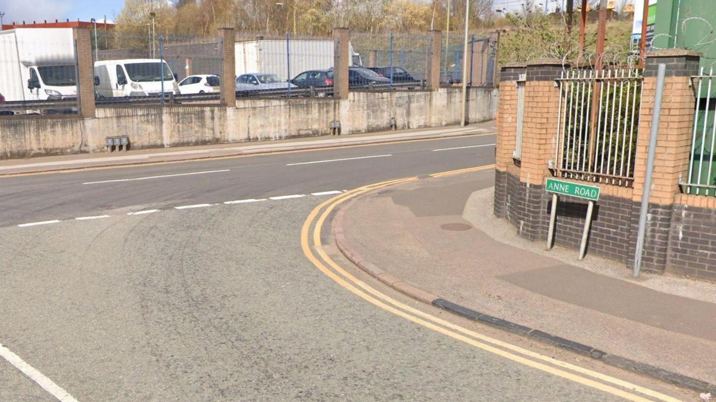 A Google street view image of the T-junction at Anne Road and Booth Street. A green sign saying Anne Road is on the right in front of a fence with brick pillars. A car park is visible behind a fence in the background.