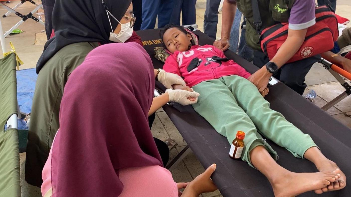 A young girl in a pink jumper and green pants lies on a foldout bed with a bottle of medicine resting against her leg while three adults sit around her, one of them holding her hand