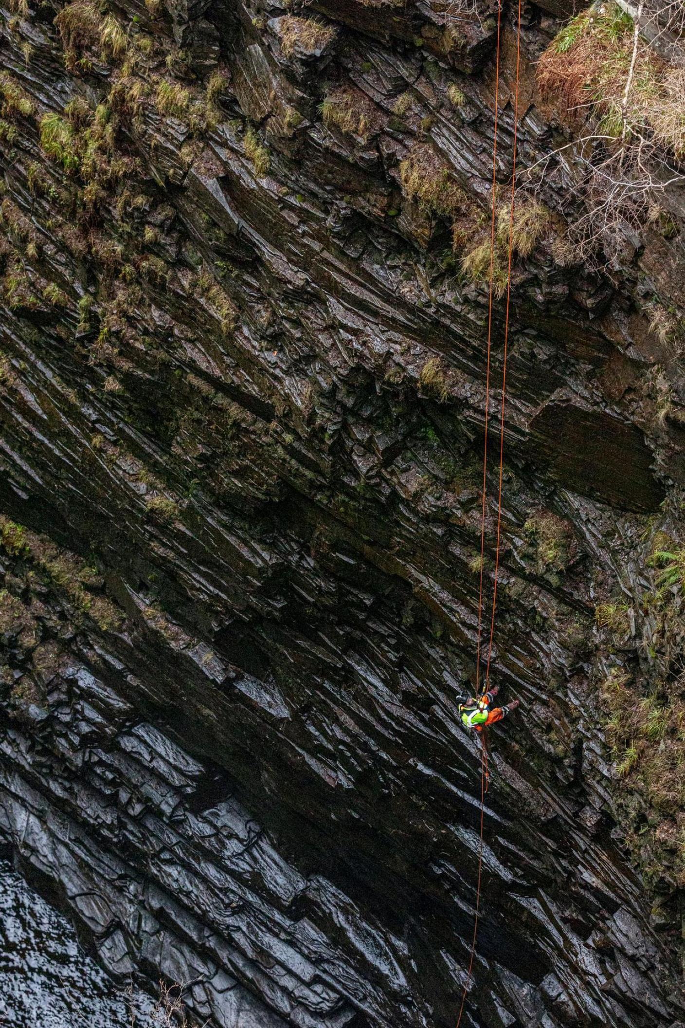 A contractor wearing protective clothing and a hard hat abseils down into the gorge. The crag's rocks are dark and wet.