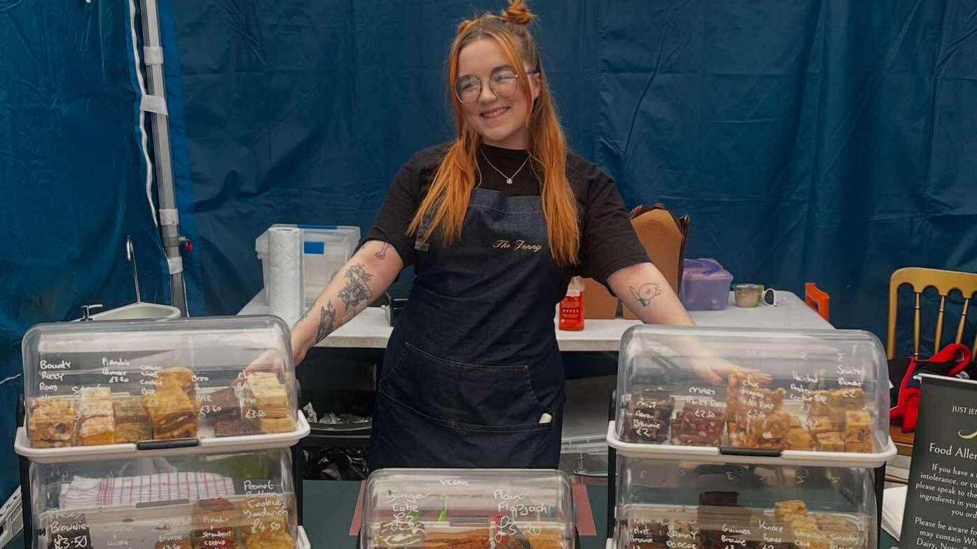 Jenny Merrick wears glasses and a dark apron. She has vibrant orange hair. She points to trays of cakes in clear boxes in a marquee with blue walls.