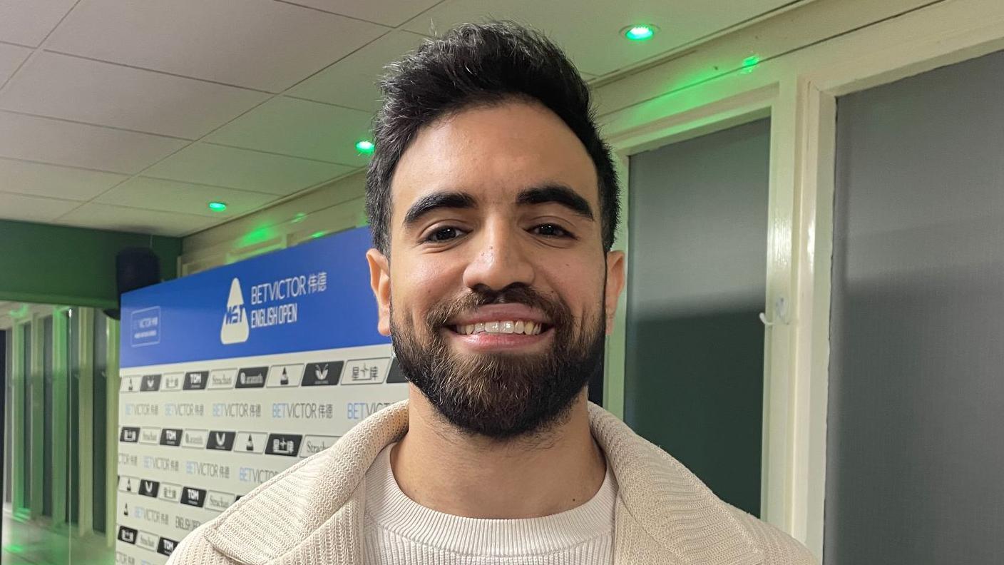 Faisal Labban has dark hair and a beard. He is smiling in front of windows with white panels and is wearing a cream jacket over a white T-shirt. Behind him is a white and blue board that has the tournament's sponsors on it.