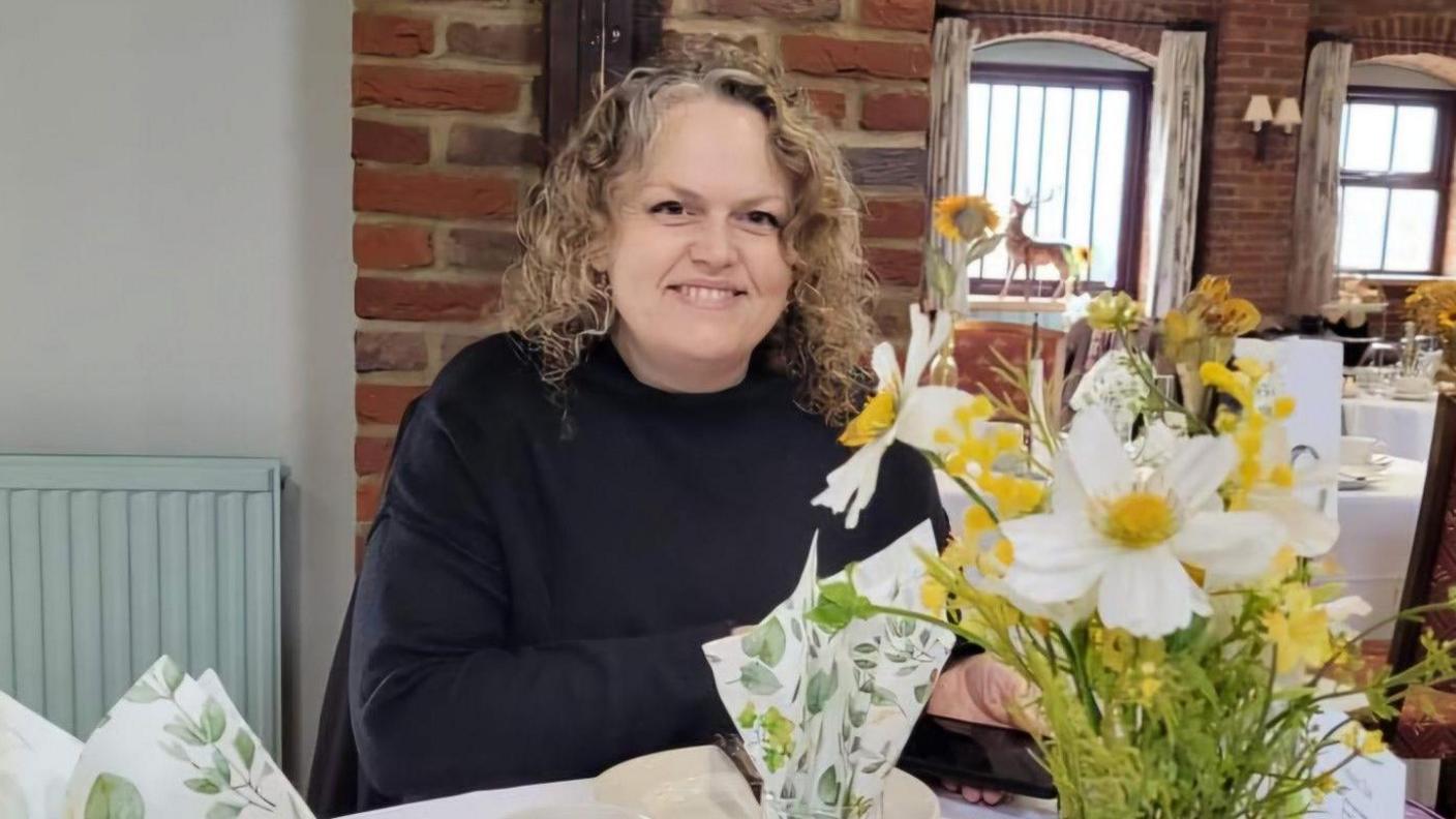 A woman sits at a dinner table in an otherwise empty restaurant. She is wearing a black jumper and has shoulder length curly hair which is light brown. There are flowers on the table in front of her that are yellow and white and a glass next to them with a serviette sticking out it covered in green leaves.