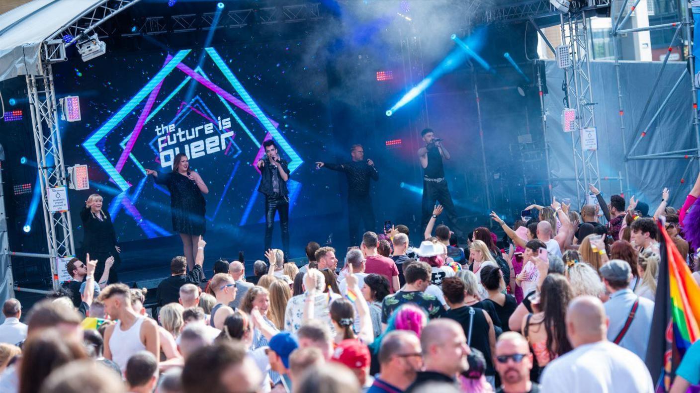 The Future is Queer performing on stage at Newcastle Pride in 2024. Michael T Ogilvie is standing on the right of the stage wearing all black and singing. He is joined on stage by four other singers. Some of the audience wave their arms in support.