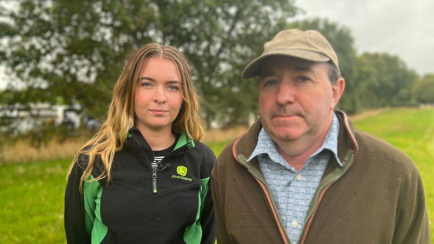 Fleur and Mark Orlik looking at the camera standing in an agricultural field used for grazing horses with trees on the background. Fleur is on the left and Mark is on the right. Fleur is wearing a green and black John Deere jacket, Mark is wearing a blue button shirt with a brown jacket and a beige cap. 