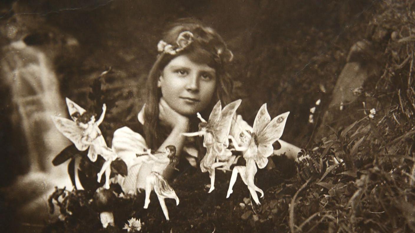 A black and white photo of a young girl with brown hair and a headband resting her hand on her neck and looking at the camera. In front of her are small dancing fairies. She is sitting outdoors with what appears to be a stream behind her.