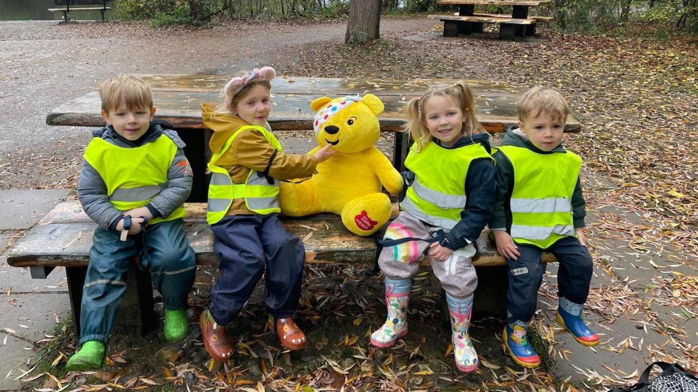 Four young children in hi-vis vests sitting at a picnic bench in the park with a Pudsey teddy bear
