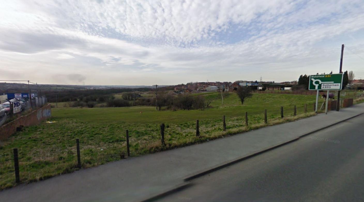 A view from an A road in Barnsley, with the road approaching a roundabout with signs towards Rotherham and Wakefield. A wire fence runs alongside the road, with a green field stretching into the distance.   