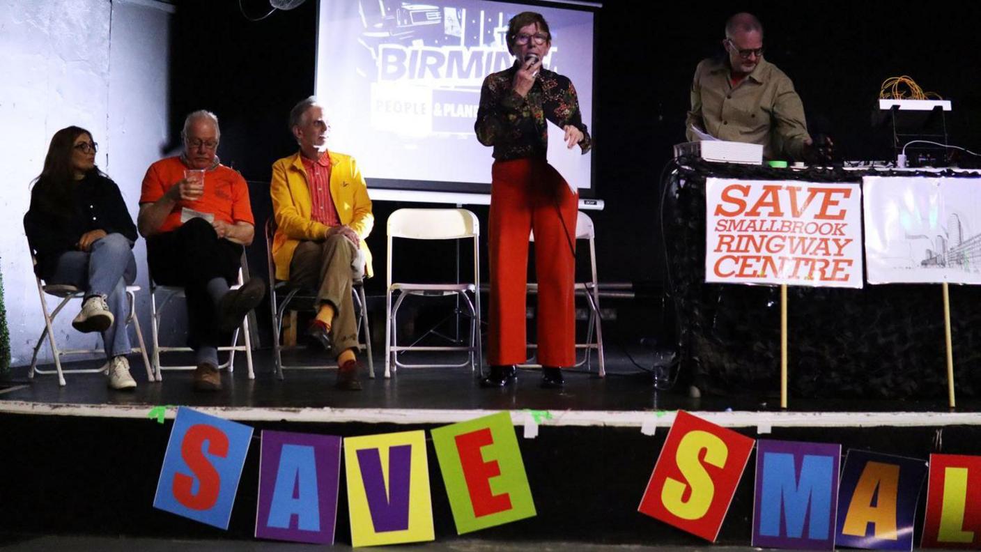 A woman and two men are sat down next to a woman in the middle, who is standing and speaking on a microphone. To her right, another man stands in front of a music system and signs.